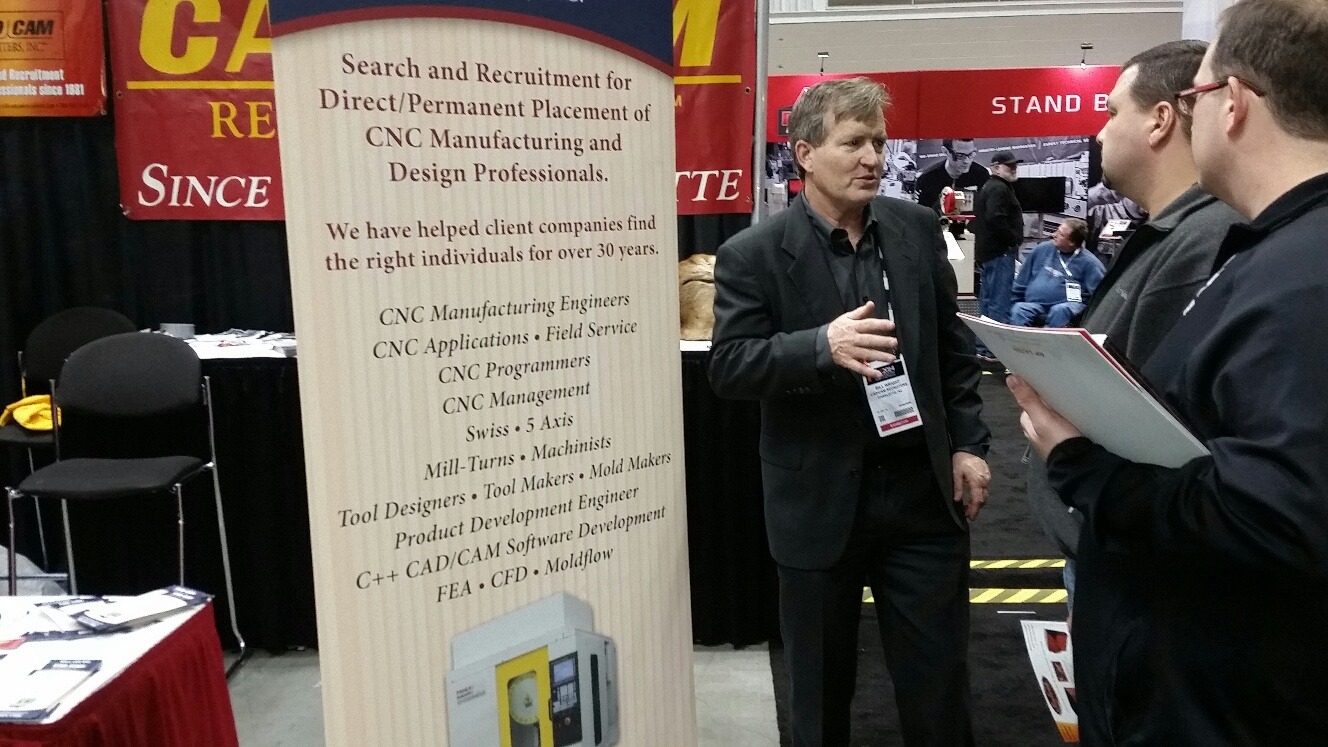 A man in a black jacket speaks to two attendees at a recruitment booth focused on CNC manufacturing and design professionals.