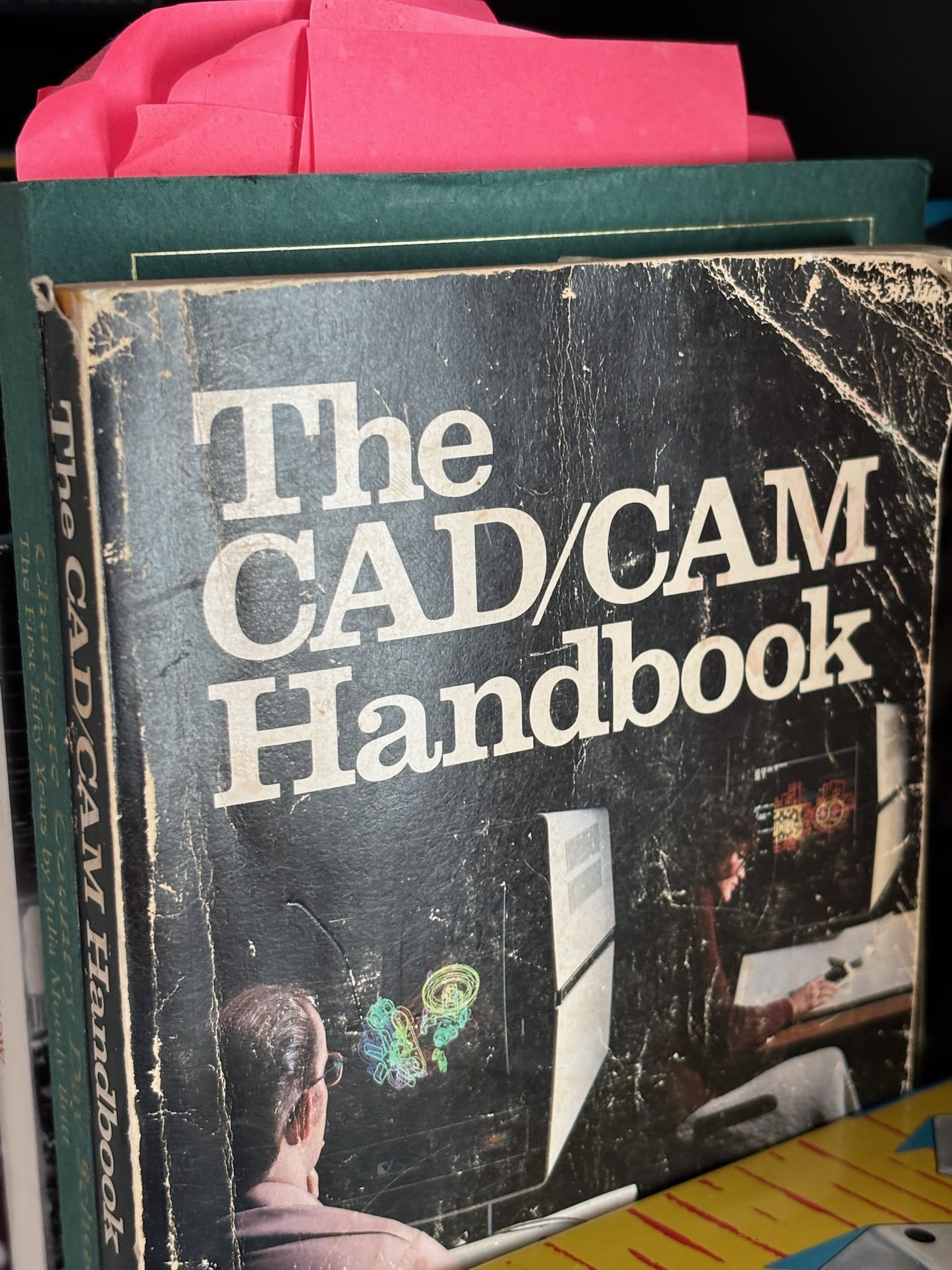The CAD/CAM Handbook, featuring a well-worn cover, shows two people using early computer-aided design technology.